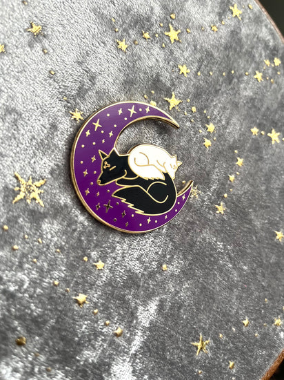 An enamel pin of the black and white dogs sleeping on a crescent moon