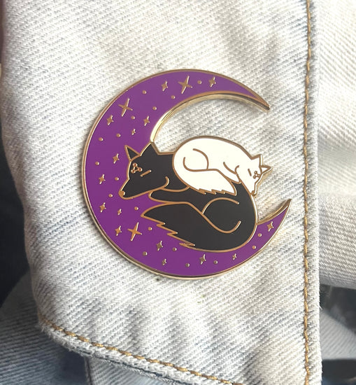 enamel pin of a purple crescent moon with a black and a white dog sleeping on it