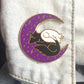 enamel pin of a purple crescent moon with a black and a white dog sleeping on it